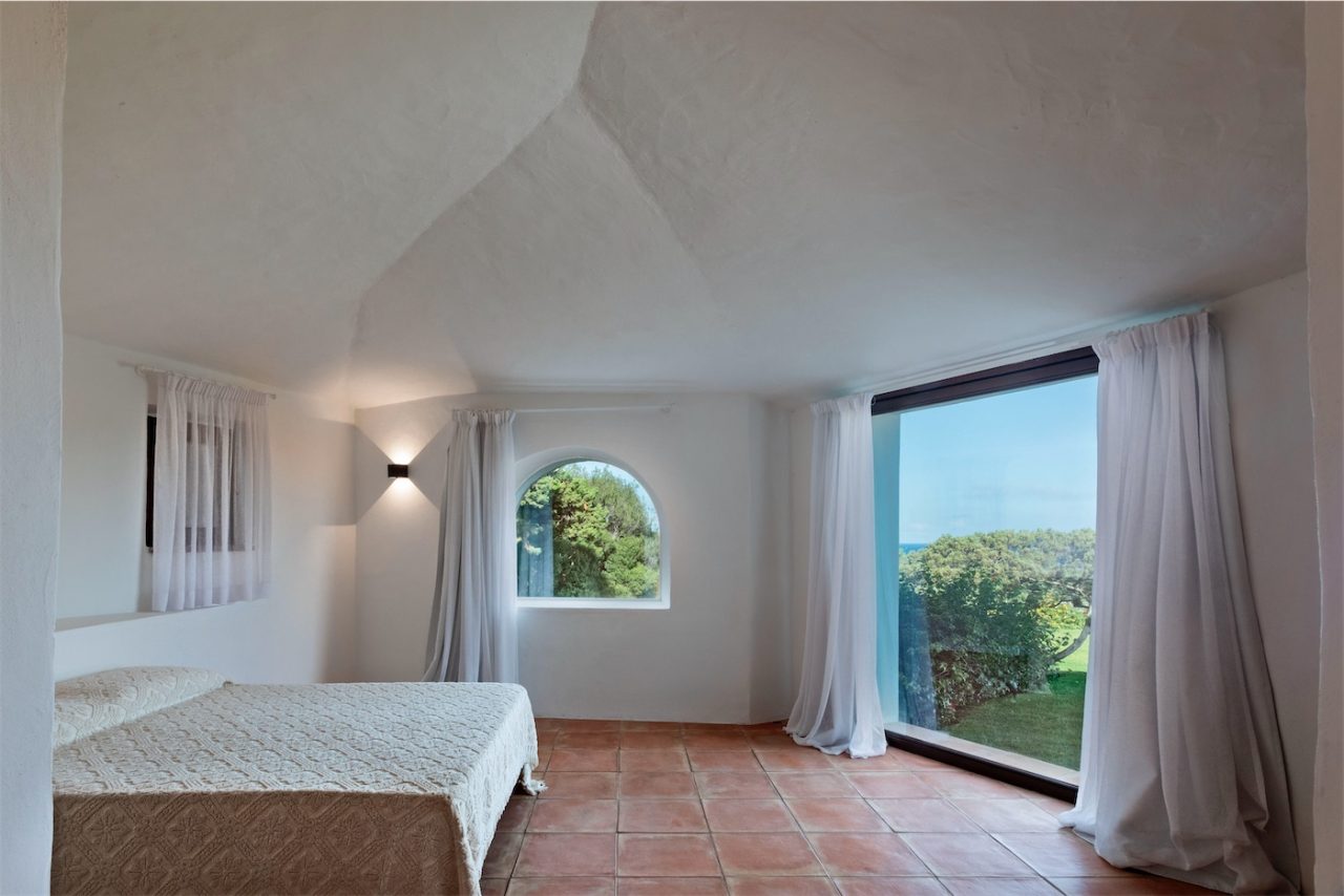 Una camera matrimoniale con un'ampia vetrata che si affaccia sul giardino. Le pareti e la stanza sono intonacate in bianco, nel classico stile della Costa Smeralda. Tende bianche in lino completano gli infissi.